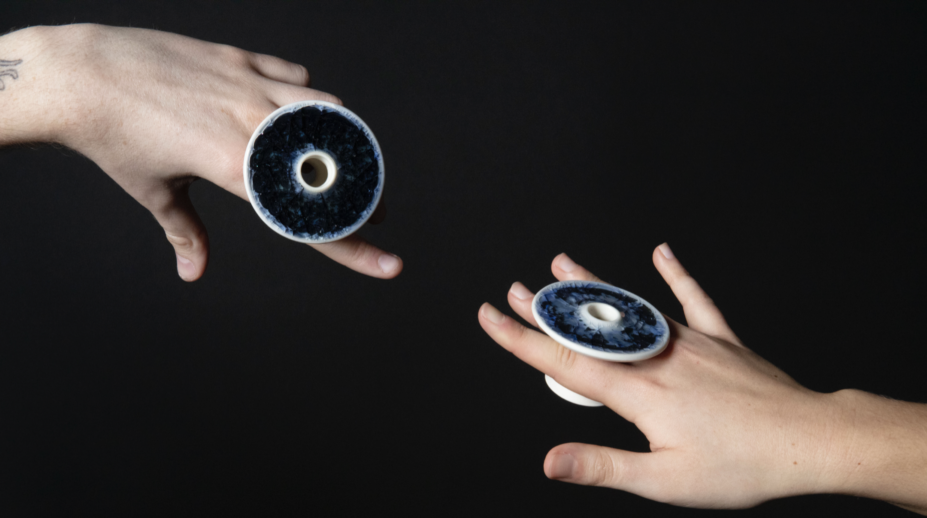 An image that includes a black background with two hands reaching out to touch from the top left corner and bottom right hand corner. Each hand holds a porcelain statement ring by Zach Dunlap between index finger and third finger.