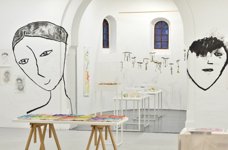Installation image of Connect / Cysylltu exhibition. Various artworks shown on a table within the apse with two faces painted directly onto white walls at each side of apse entrance