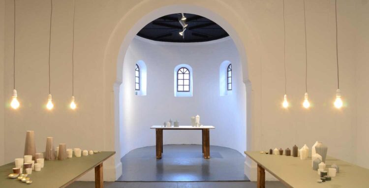 Shifting Lines exhibition by Justine Allison, part of The Language of Clay programme. Three tables painted with different tones of green, two at forefront and one within the apse. Each has a collection of the artist's pieces on top, with three hanging bulbs lighting the pieces on each table.